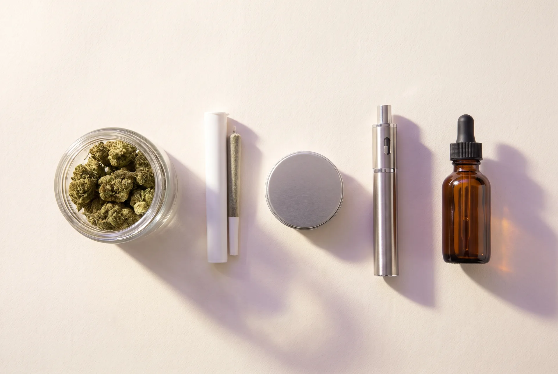 Overhead arrangement of the cannabis product formats Purple Buds carries: cured flower in a glass jar, a pre-roll, an edible tin, a vape pen, and a tincture dropper bottle