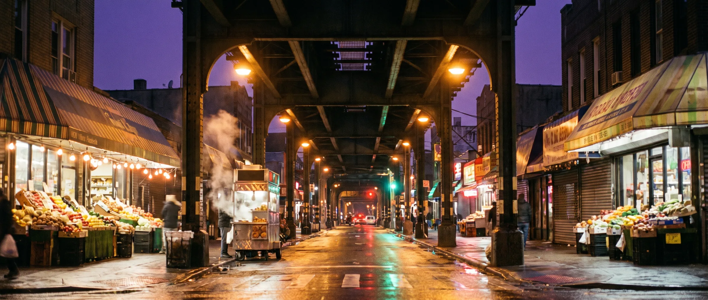 Roosevelt Avenue at night under the elevated 7 train in Jackson Heights, Queens, awnings and market stalls lit by sodium lamps on the block Purple Buds Dispensary sits on